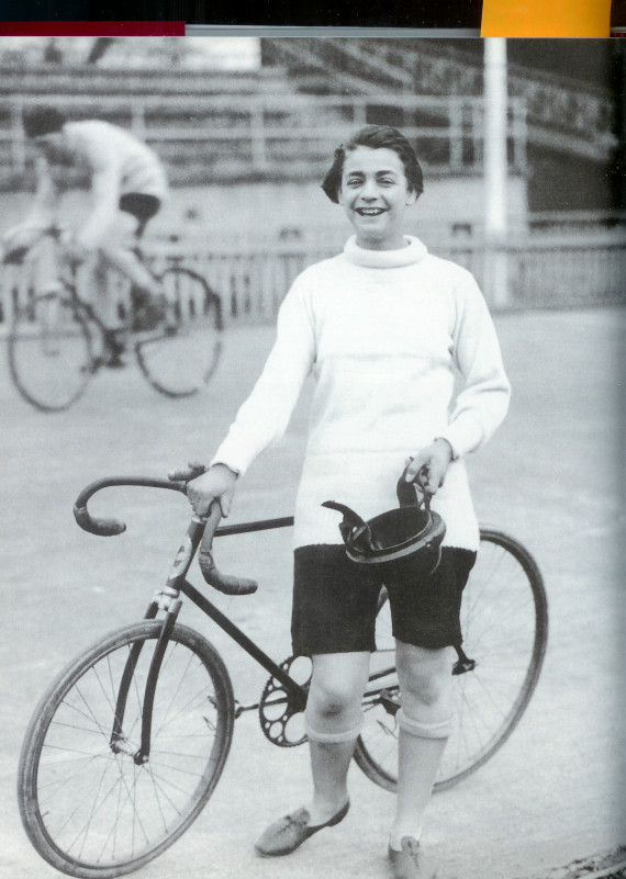 Im Vordergrund steht ein junger Mann mit seinem Rennrad, in der Hand den Helm, er lacht und freut sich möglicherweise über seinen Sieg. Im Hintergrund fährt ein anderer Mann auf einem Rennrad.